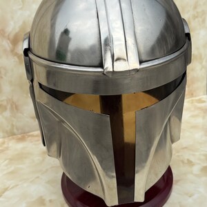 Puede incluir: Un casco plateado con una visera en forma de T, que recuerda a un casco mandaloriano. El casco se exhibe sobre un soporte rojo oscuro, mostrando su dise&ntilde;o detallado y su artesan&iacute;a. La superficie del casco refleja la luz.