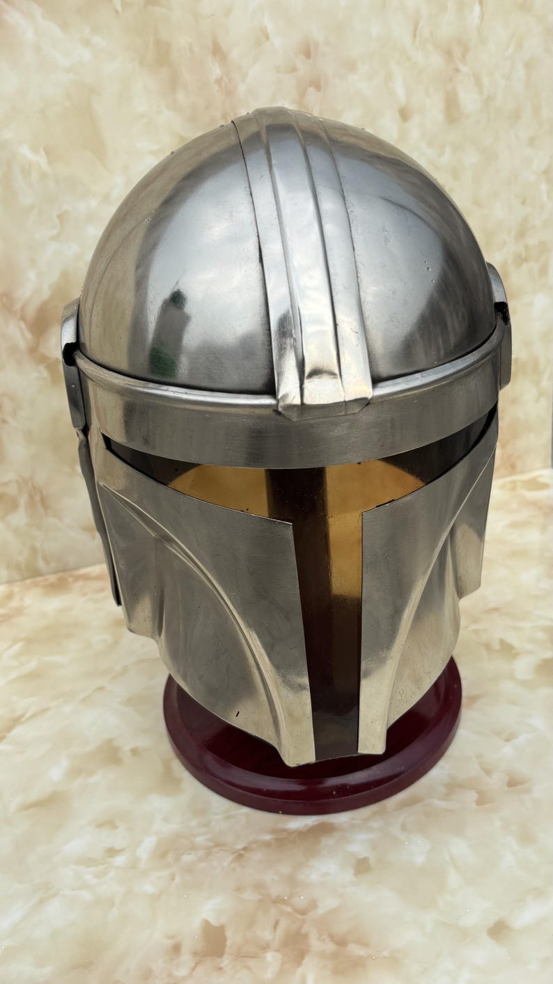 Puede incluir: Un casco plateado con una visera en forma de T, que recuerda a un casco mandaloriano. El casco se exhibe sobre un soporte rojo oscuro, mostrando su dise&ntilde;o detallado y su artesan&iacute;a. La superficie del casco refleja la luz.