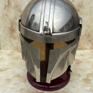 Puede incluir: Un casco plateado con visera dorada, sobre una base roja oscura. El casco tiene crestas verticales y un dise&ntilde;o angular distintivo. El fondo presenta un patr&oacute;n veteado.