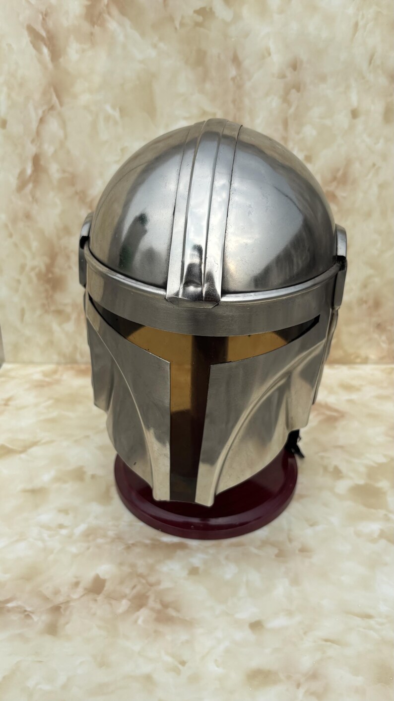 Puede incluir: Un casco plateado con visera dorada, sobre una base roja oscura. El casco tiene crestas verticales y un dise&ntilde;o angular distintivo. El fondo presenta un patr&oacute;n veteado.