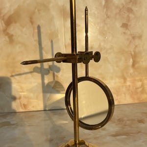 May include: A brass-colored, vintage-style magnifying glass with a circular lens and adjustable arm. The base is round with concentric rings. The design includes a pointed tip and a small, vertical post.