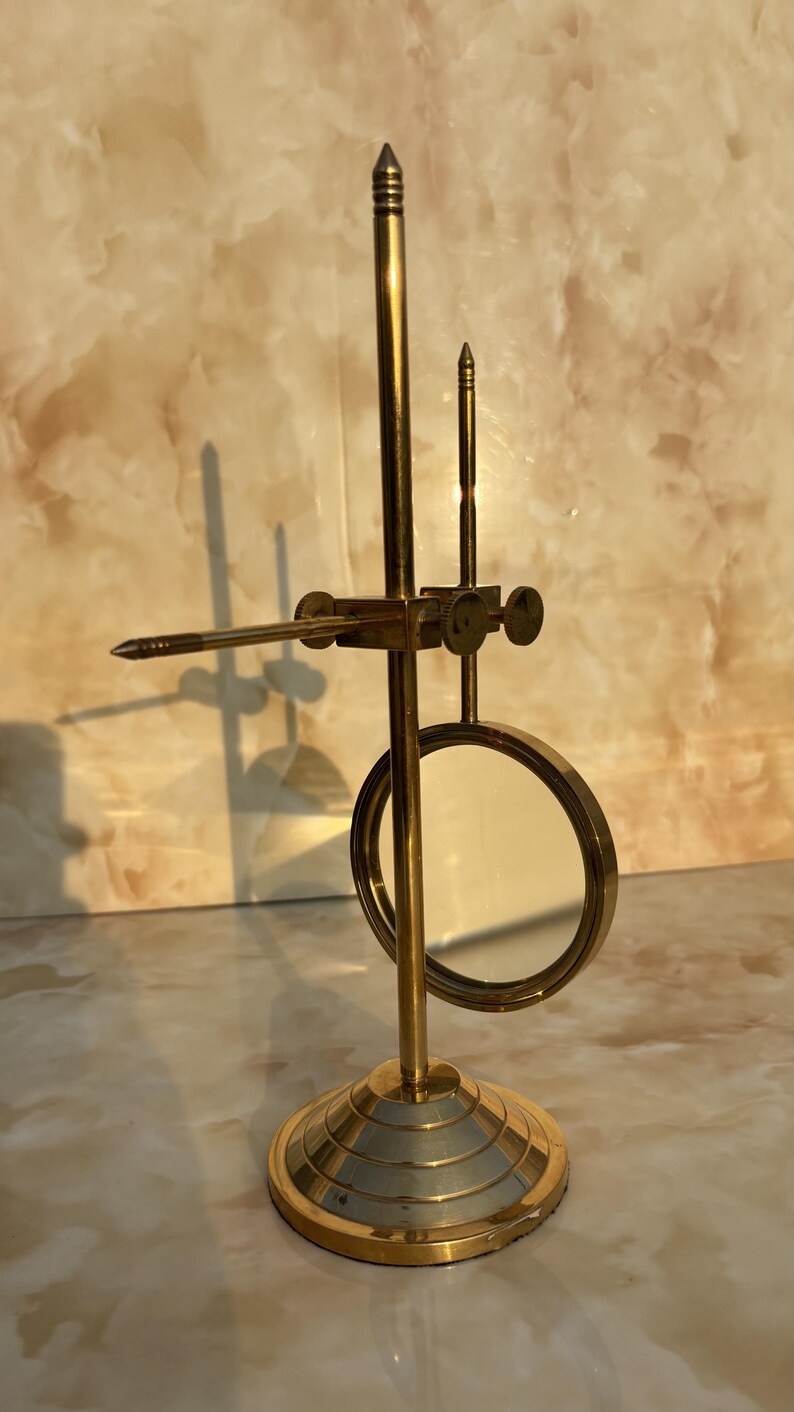 May include: A brass-colored, vintage-style magnifying glass with a circular lens and adjustable arm. The base is round with concentric rings. The design includes a pointed tip and a small, vertical post.