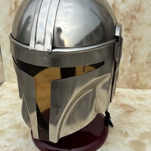 Puede incluir: Un casco met&aacute;lico plateado con visera dorada, sobre un soporte rojo oscuro. El casco tiene una parte superior redondeada con crestas verticales y una visera rectangular. Probablemente sea un accesorio o parte de un disfraz.