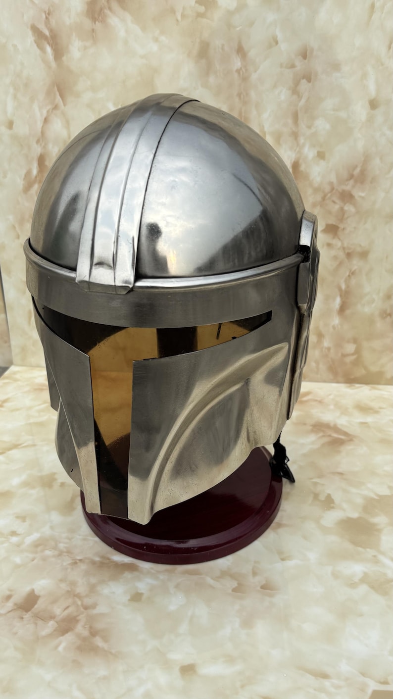 Puede incluir: Un casco met&aacute;lico plateado con visera dorada, sobre un soporte rojo oscuro. El casco tiene una parte superior redondeada con crestas verticales y una visera rectangular. Probablemente sea un accesorio o parte de un disfraz.