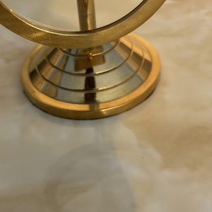 May include: A gold-colored, round vanity mirror with a magnifying glass. The mirror has a circular frame and a pedestal base. The base has a tiered design. The mirror is on a light-colored surface.