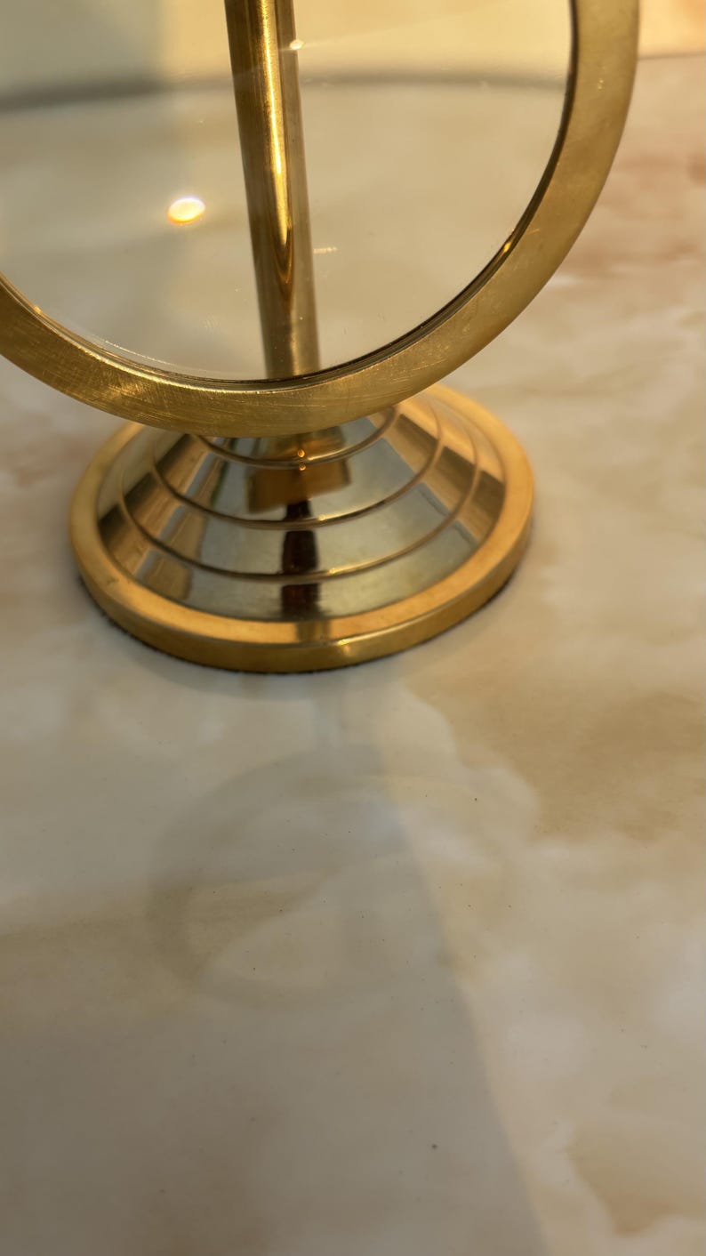 May include: A gold-colored, round vanity mirror with a magnifying glass. The mirror has a circular frame and a pedestal base. The base has a tiered design. The mirror is on a light-colored surface.
