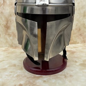 Puede incluir: Un casco plateado met&aacute;lico con una distintiva visera en forma de T, apoyado sobre un soporte rojo oscuro. El dise&ntilde;o del casco presenta una parte superior redondeada con crestas verticales y placas angulares en las mejillas, lo que sugiere un tema futurista o de ciencia ficci&oacute;n.