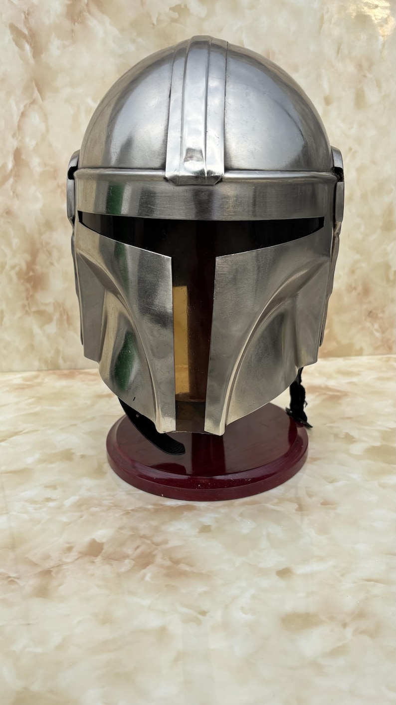 Puede incluir: Un casco plateado met&aacute;lico con una distintiva visera en forma de T, apoyado sobre un soporte rojo oscuro. El dise&ntilde;o del casco presenta una parte superior redondeada con crestas verticales y placas angulares en las mejillas, lo que sugiere un tema futurista o de ciencia ficci&oacute;n.