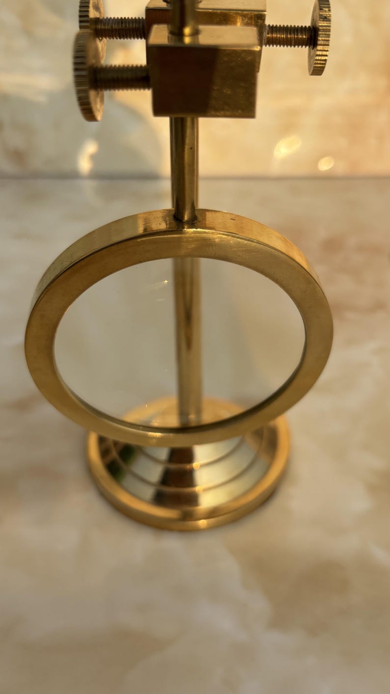 May include: A vintage-style magnifying glass with a brass finish. The round lens is held in place by a brass frame, and the base has a tiered design. The adjustable focus mechanism is visible at the top. This desk accessory is ideal for reading or detailed work.