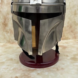 Puede incluir: Casco mandaloriano plateado con visor en forma de T, sobre un soporte rojo oscuro. El casco muestra un acabado met&aacute;lico y un dise&ntilde;o detallado. Un art&iacute;culo de colecci&oacute;n.