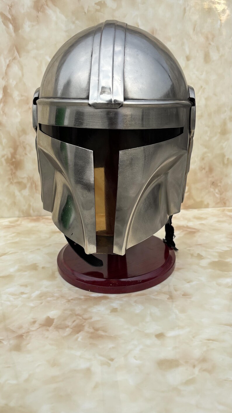 Puede incluir: Casco mandaloriano plateado con visor en forma de T, sobre un soporte rojo oscuro. El casco muestra un acabado met&aacute;lico y un dise&ntilde;o detallado. Un art&iacute;culo de colecci&oacute;n.