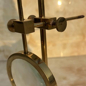 May include: A vintage brass magnifying glass with a round lens, mounted on a stand with adjustable arms and a circular base. The brass has a warm, golden hue, and the lens is clear. The design suggests a scientific or antique aesthetic.