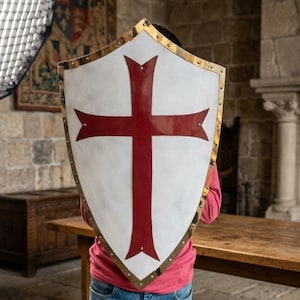 Könnte beinhalten: Ein weißer Schild mit einem roten Kreuz-Design und goldenen Verzierungen. Der Schild wird von einer Person getragen, die ein rotes Hemd und blaue Jeans trägt. Im Hintergrund ein Holztisch und Steinmauern.