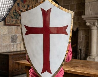 May include: A white shield with a red cross design and gold trim. The shield is held in front of a person wearing a red shirt and blue jeans. The background includes a wooden table and stone walls.