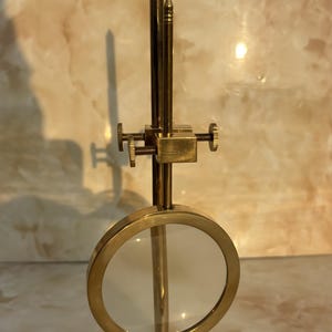 May include: A brass-colored magnifying glass with a round lens and a detailed base. The magnifying glass has a vertical support with adjustable components and a small, pointed top. The design is reminiscent of antique scientific instruments.