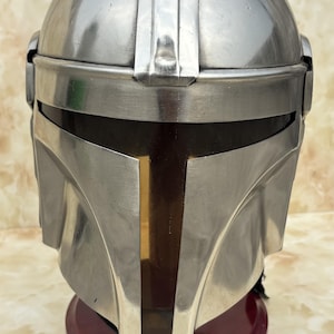 Puede incluir: Un casco mandaloriano plateado con una visera oscura y una franja vertical dorada en el centro. El casco se exhibe sobre un soporte rojo oscuro, mostrando su dise&ntilde;o detallado y acabado met&aacute;lico.