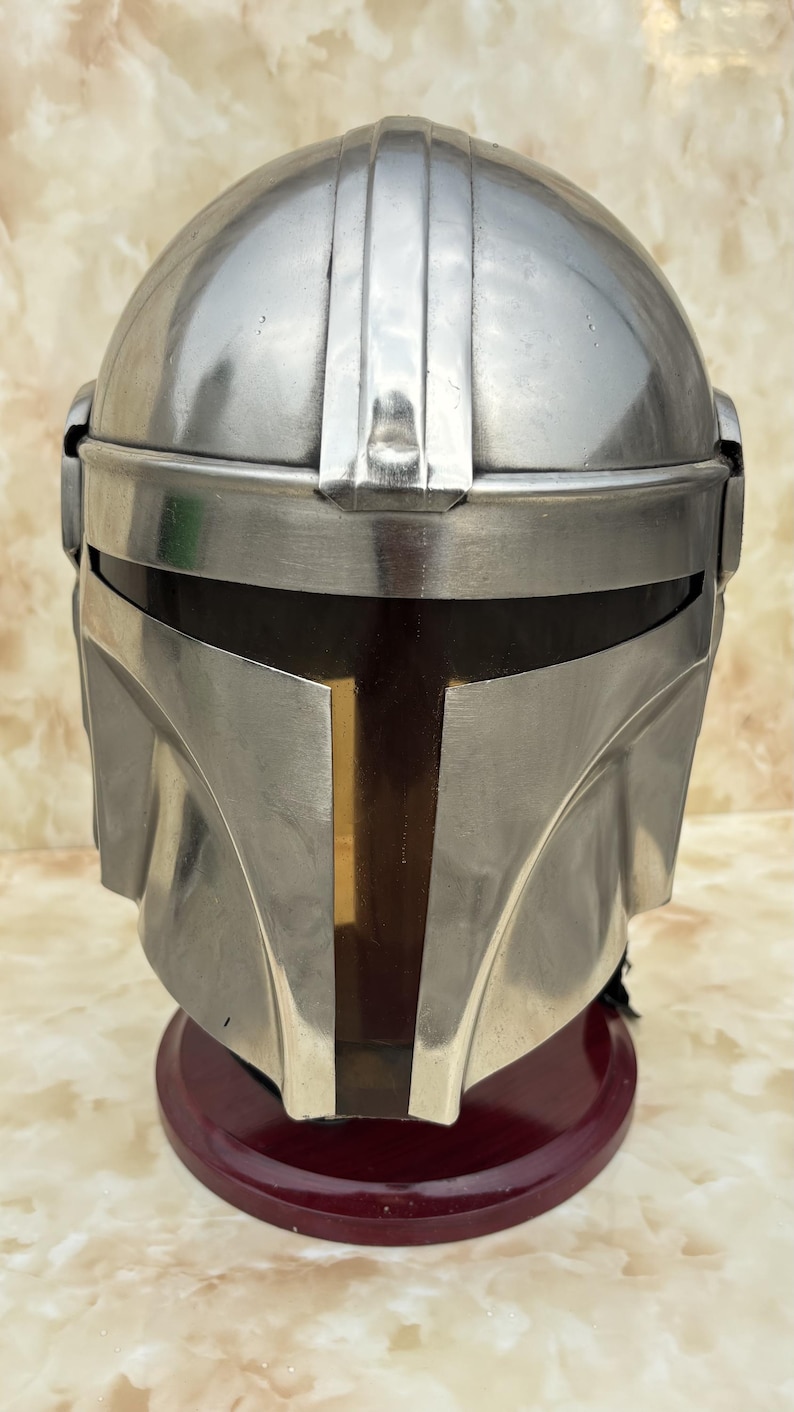 Puede incluir: Un casco mandaloriano plateado con una visera oscura y una franja vertical dorada en el centro. El casco se exhibe sobre un soporte rojo oscuro, mostrando su dise&ntilde;o detallado y acabado met&aacute;lico.