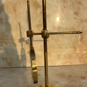 May include: A brass-colored scientific instrument with a circular magnifying lens and adjustable components. The base is circular with concentric rings. The instrument has a vertical rod and a horizontal arm with a pointed tip.