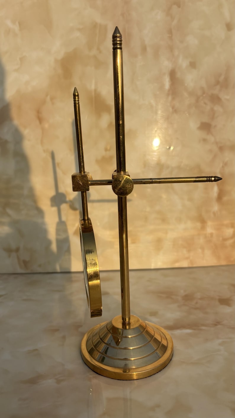 May include: A brass-colored scientific instrument with a circular magnifying lens and adjustable components. The base is circular with concentric rings. The instrument has a vertical rod and a horizontal arm with a pointed tip.