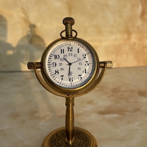 Pode incluir: Um relógio de mesa de latão em estilo vintage, com mostrador redondo e algarismos romanos. O relógio tem um design detalhado e assenta numa base decorativa. O mostrador do relógio exibe os números de 1 a 12, com números adicionais.