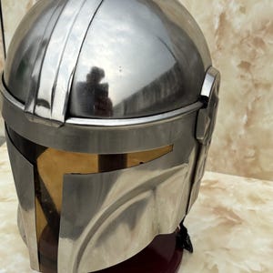 Puede incluir: Un casco met&aacute;lico plateado con visera, que recuerda a un casco mandaloriano. El casco tiene una parte superior redondeada con una cresta central y una visera oscura. El casco se exhibe sobre un soporte rojo oscuro.
