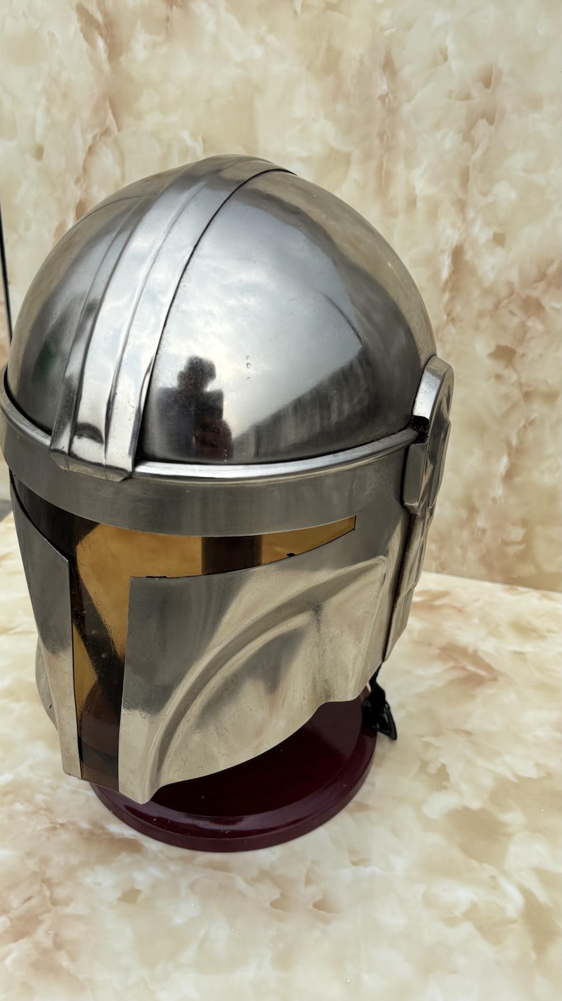 Puede incluir: Un casco met&aacute;lico plateado con visera, que recuerda a un casco mandaloriano. El casco tiene una parte superior redondeada con una cresta central y una visera oscura. El casco se exhibe sobre un soporte rojo oscuro.