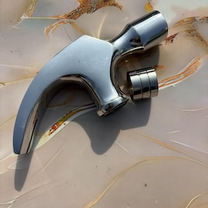 Könnte beinhalten: Ein polierter Chromhammer mit gebogenem Griff und zylindrischem Kopf. Der Hammer hat eine glatte, reflektierende Oberfläche und ist zum Schlagen konzipiert. Ein Satz von drei Ringen ist am Hammer befestigt.