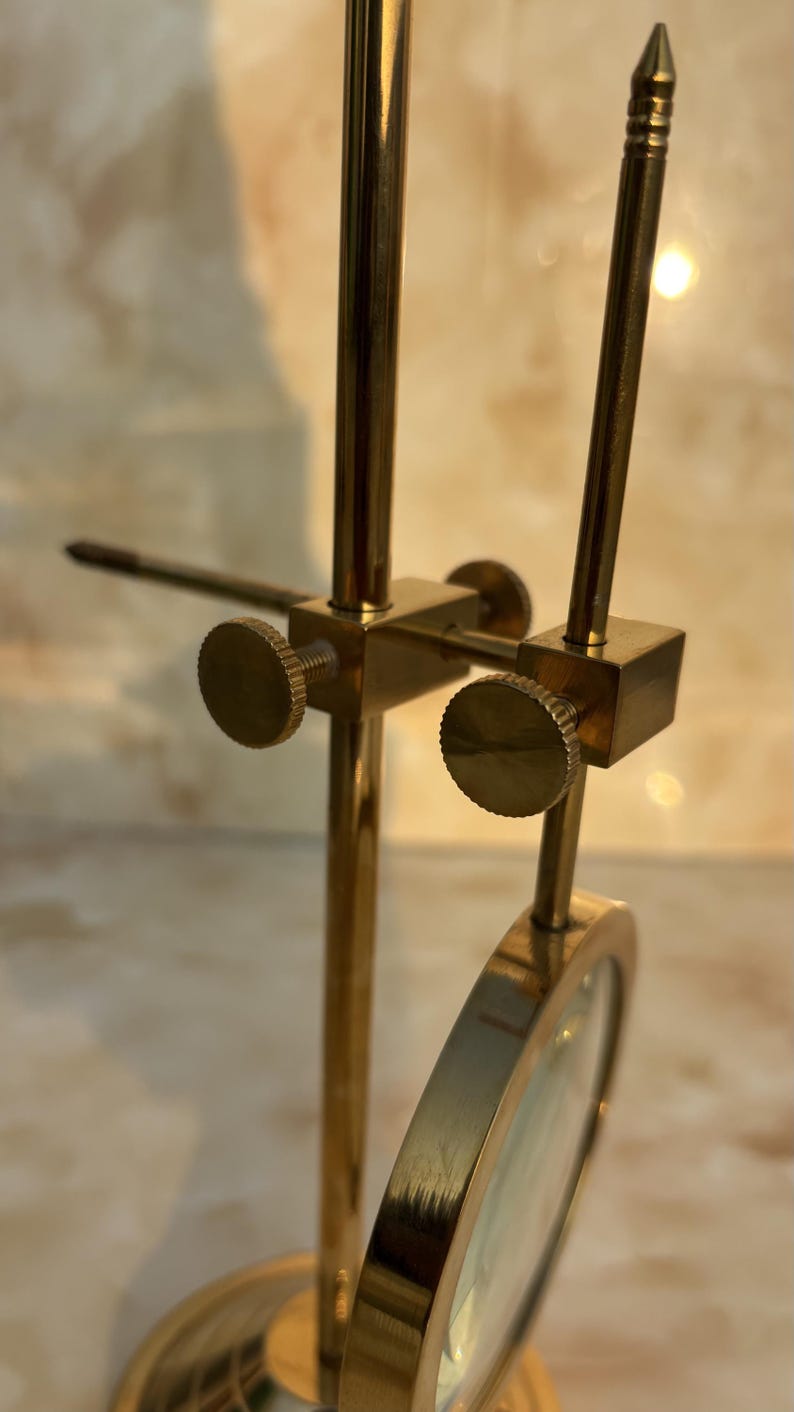 May include: A vintage brass magnifying glass with a round lens and adjustable stand. The stand features a circular base, vertical support, and adjustable arms with knurled knobs. The magnifying glass is designed for detailed viewing.