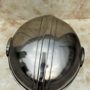 Puede incluir: Un objeto redondeado y brillante de metal con ranuras verticales. El objeto tiene forma circular con una parte superior ligeramente aplanada y dos peque&ntilde;as protuberancias redondas a cada lado. La superficie refleja la luz.