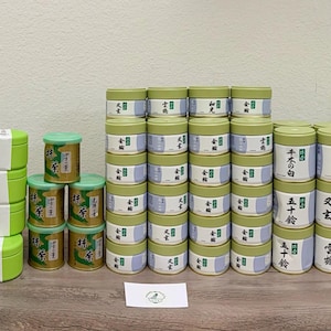 Op de afbeelding: Een verzameling kleine, ronde theeblikken in verschillende kleuren, waaronder groen, paars en goud, gerangschikt op een houten oppervlak. De blikken hebben groene deksels en witte etiketten met Japanse tekst.