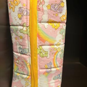May include: A rectangular, quilted fabric case with a yellow zipper. The case is pink with a repeating pattern of cartoon bears, stars, and rainbows. The zipper runs vertically down the center of the case.
