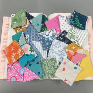 May include: A collection of colorful fabric squares, each with unique patterns and designs, arranged on an open book. The squares feature floral, geometric, and abstract prints in shades of pink, blue, green, and orange. The book's pages are visible beneath the fabric.