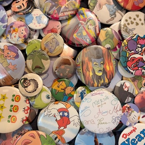 Puede incluir: Una colección de coloridos botones redondos con varios personajes y diseños de dibujos animados. Algunos botones tienen texto, incluyendo "Disney's Yearbook" y firmas. Los botones varían en tamaño y color, creando un surtido vibrante.