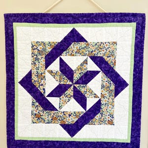 May include: A square wall hanging quilt featuring a central star pattern with purple fabric and a floral print of white, orange, and green flowers. The quilt has a light green inner border and a wide purple outer border with a white rope hanger.