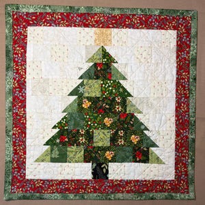 May include: A square quilted wall hanging featuring a central Christmas tree design composed of various green and floral patterned fabric patches. The tree is set against a white background with a red border and a green outer edge with leaf motifs.