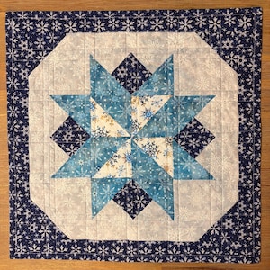 May include: A square quilted wall hanging featuring a central eight-pointed star pattern. The design incorporates shades of navy blue, light blue, and white fabric with a repeating snowflake print. The quilt is finished with a dark blue border.