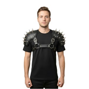 May include: Black leather shoulder armor with silver spikes. The armor features adjustable straps and metal buckles. The design includes a chest harness with metal rings. The product is designed to be worn over clothing.