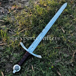 May include: A silver sword with a brown leather-wrapped handle and a cross-shaped guard. The blade has etched designs and the words "VINCIT" and "CHRISTUS". The sword rests on a bed of grass and dirt.