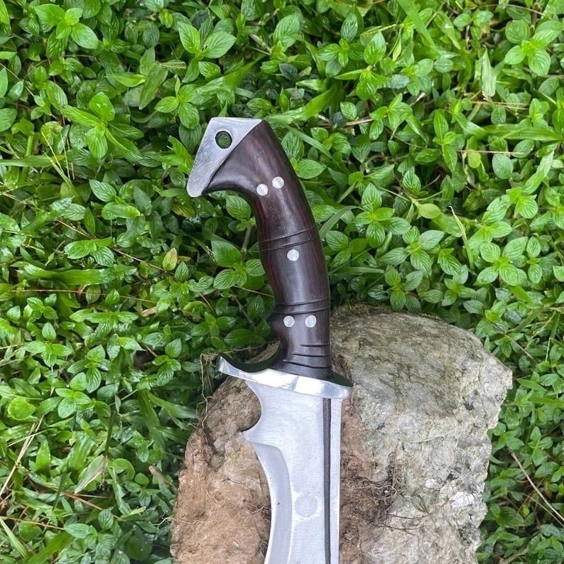 May include: A handcrafted dagger with a dark wooden handle and a silver blade. The handle features silver rivets and a curved design. The blade has a sharp edge and a pointed tip. The dagger is displayed against a backdrop of green foliage and a stone.