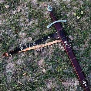 May include: A medieval-style sword with a brown leather sheath and handle, resting on the ground. The sword features a silver crossguard and a blade with the inscription "VINCIT" and "CHRISTUS". A black leather belt with white crosses is also present.