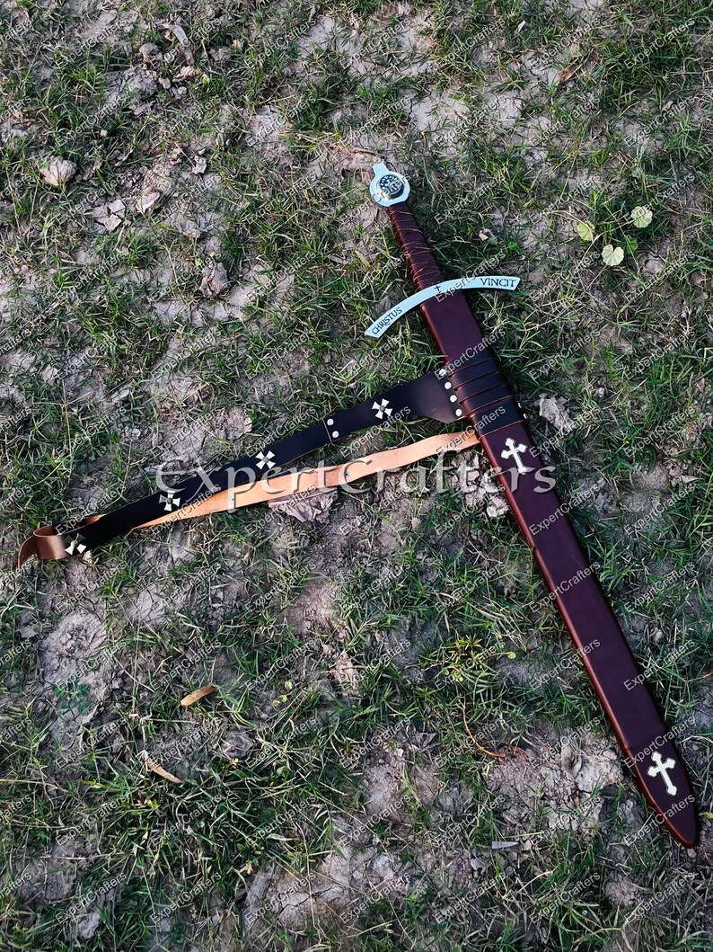 May include: A medieval-style sword with a brown leather sheath and handle, resting on the ground. The sword features a silver crossguard and a blade with the inscription "VINCIT" and "CHRISTUS". A black leather belt with white crosses is also present.