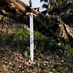 May include: A decorative sword with a silver blade and ornate gold hilt, featuring a red gem. The handle is wrapped in black material. The sword is displayed outdoors, leaning against a large tree trunk.