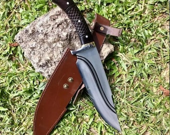 May include: A large hunting knife with a dark wooden handle and a silver blade, resting in a brown leather sheath. The knife features a textured handle and a curved blade design. The sheath has two rivets and a belt loop. The knife is set on a stone and grass background.