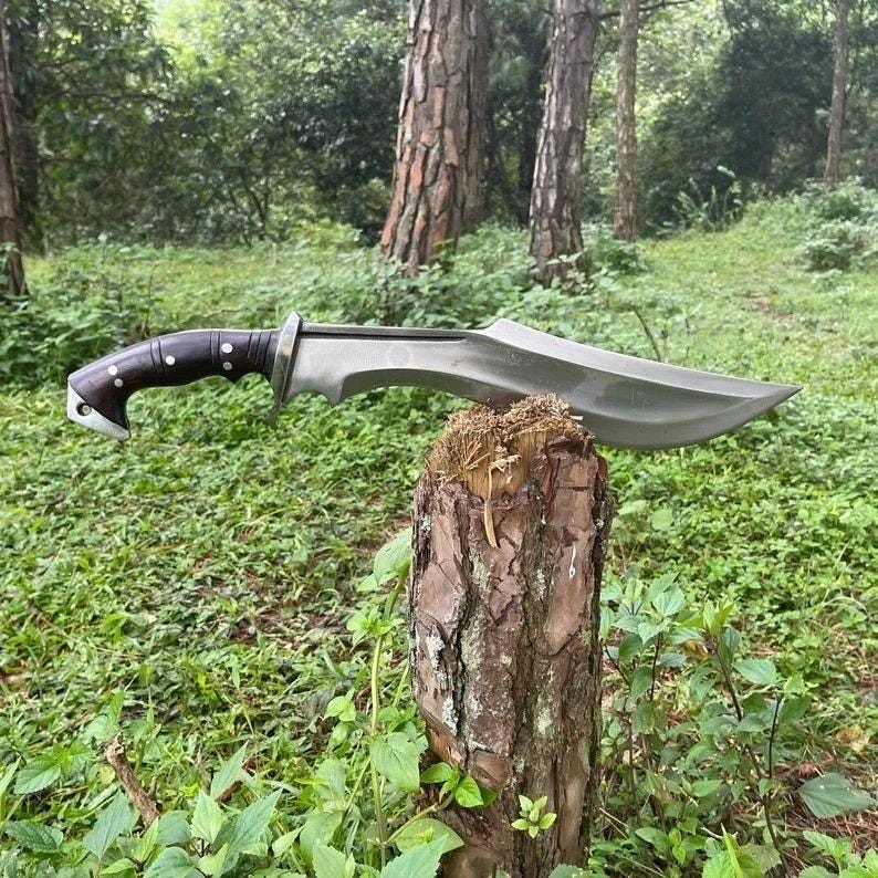 May include: A large, curved blade knife with a dark wooden handle and silver accents. The blade is a matte gray color. The knife is displayed on a tree stump in a natural outdoor setting.