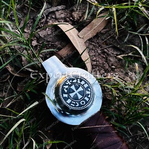 May include: A silver-colored sword hilt with a circular emblem featuring a cross design and text. The hilt is set against a backdrop of green grass and brown leaves, with a dark brown leather-wrapped handle.