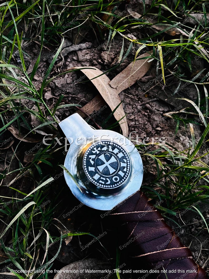 May include: A silver-colored sword hilt with a circular emblem featuring a cross design and text. The hilt is set against a backdrop of green grass and brown leaves, with a dark brown leather-wrapped handle.