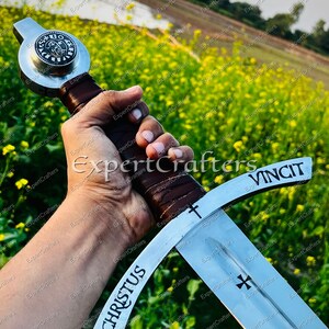 May include: A medieval sword with a silver blade and a brown leather-wrapped handle. The blade features the words "CHRISTUS" and "VINCIT", along with a cross symbol. The sword's hilt has a circular emblem.