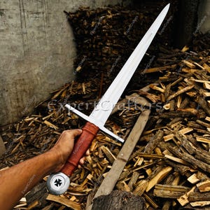 May include: A medieval sword with a long, silver blade and a cross-shaped hilt. The handle is wrapped in brown leather. The sword is held in a person's hand against a backdrop of firewood.