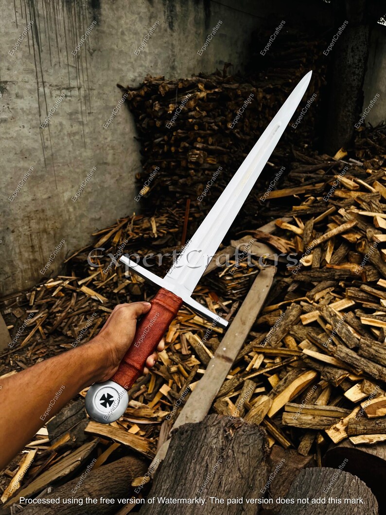 May include: A medieval sword with a long, silver blade and a cross-shaped hilt. The handle is wrapped in brown leather. The sword is held in a person's hand against a backdrop of firewood.