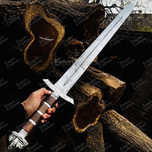 Op de afbeelding: Een lang zwaard met een patroon van staal en een houten handvat. De handgreep heeft zilveren accenten en een decoratieve pommel. Het zwaard wordt getoond tegen een achtergrond van grote, gezaagde boomstammen.